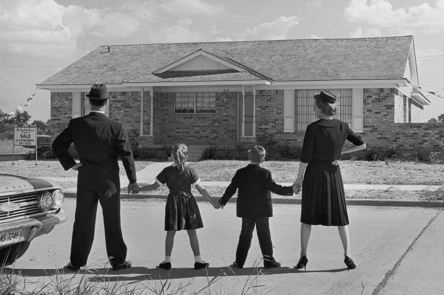 Imagem de uma família de quatro pessoas de mãos dadas em uma fila, de costas para a câmera, de frente para uma casa em estilo rancho em um empreendimento suburbano. Uma placa de venda está afixada no jardim da frente da casa.