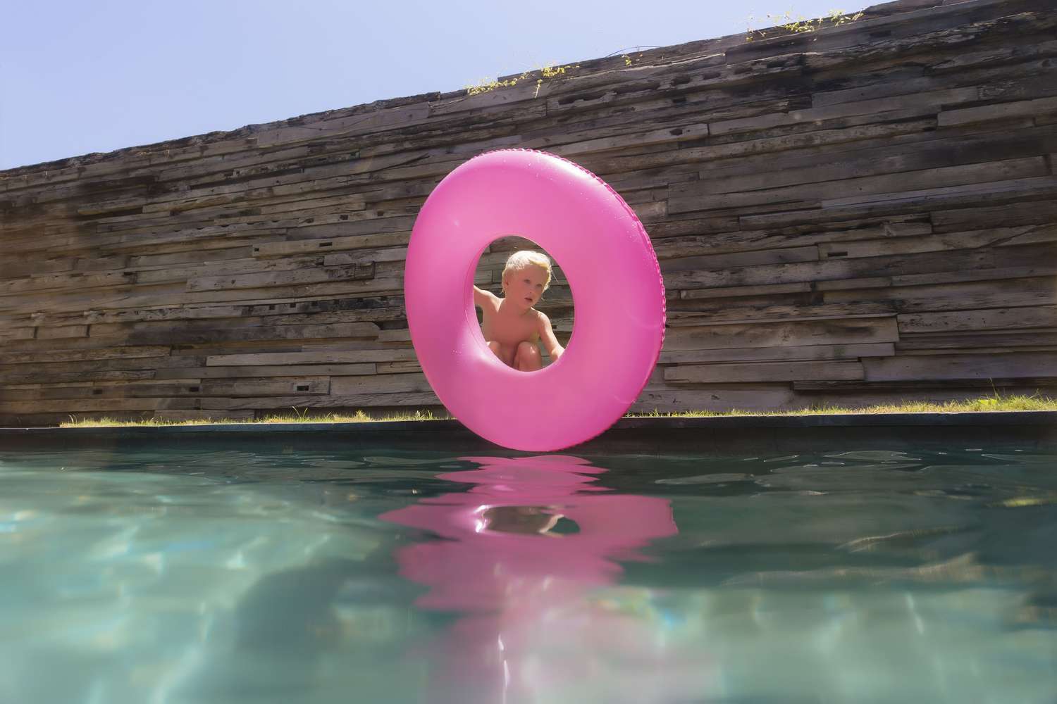 Um menino segurando uma boia de tubo de piscina rosa ao lado de uma piscina cercada por um muro de pedra.
