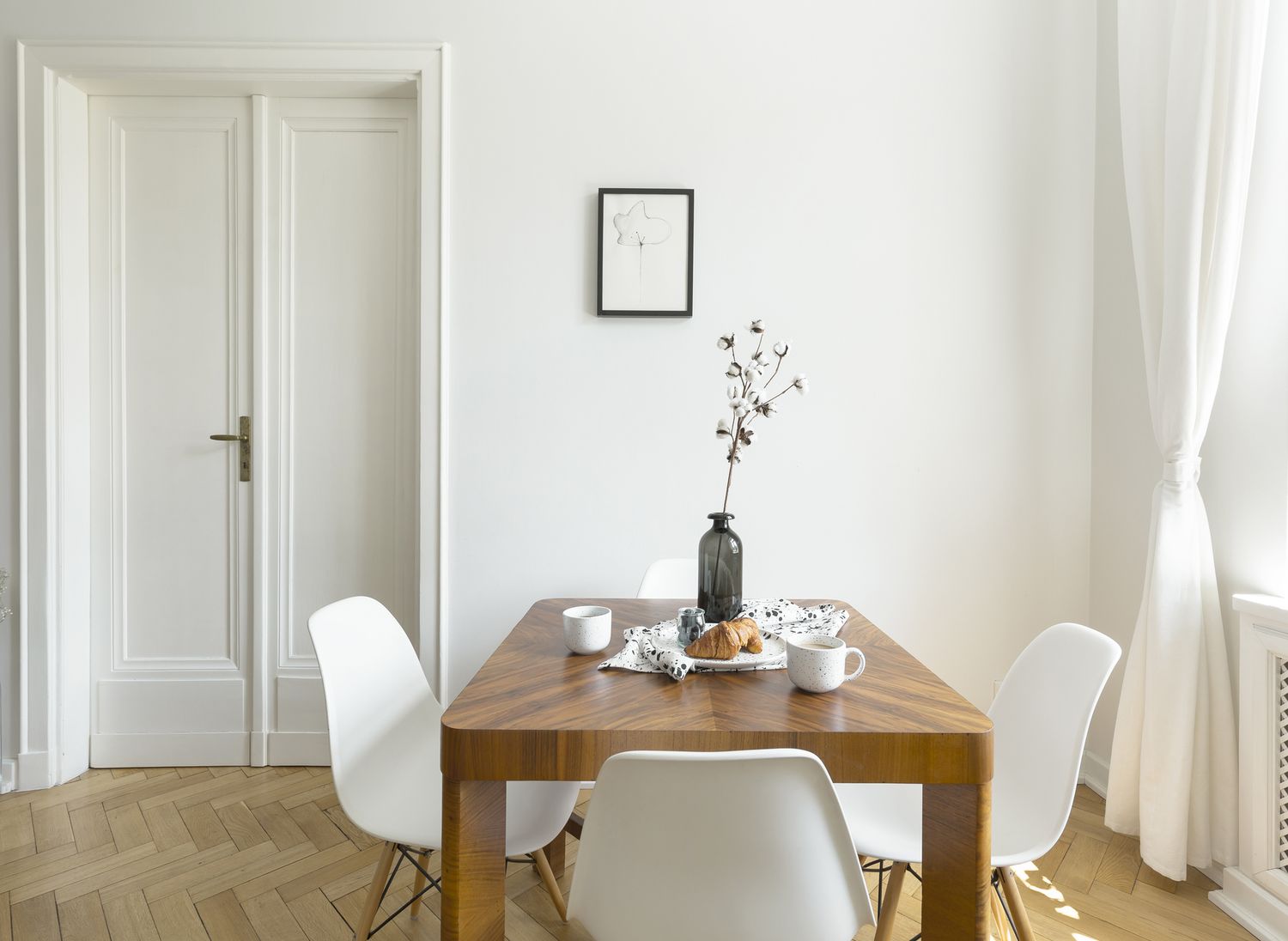 Cadeiras brancas em uma mesa de madeira no interior de uma sala de jantar minimalista com porta e pôster.