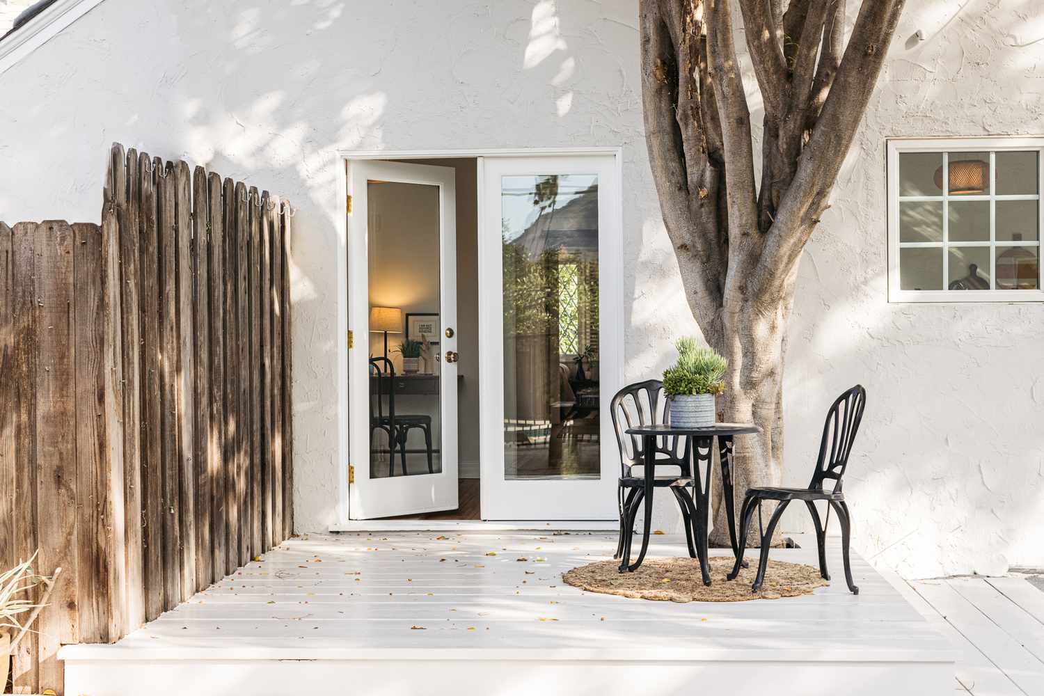 Cubierta exterior de madera blanca decorada con una mesita negra y sillas junto a un árbol