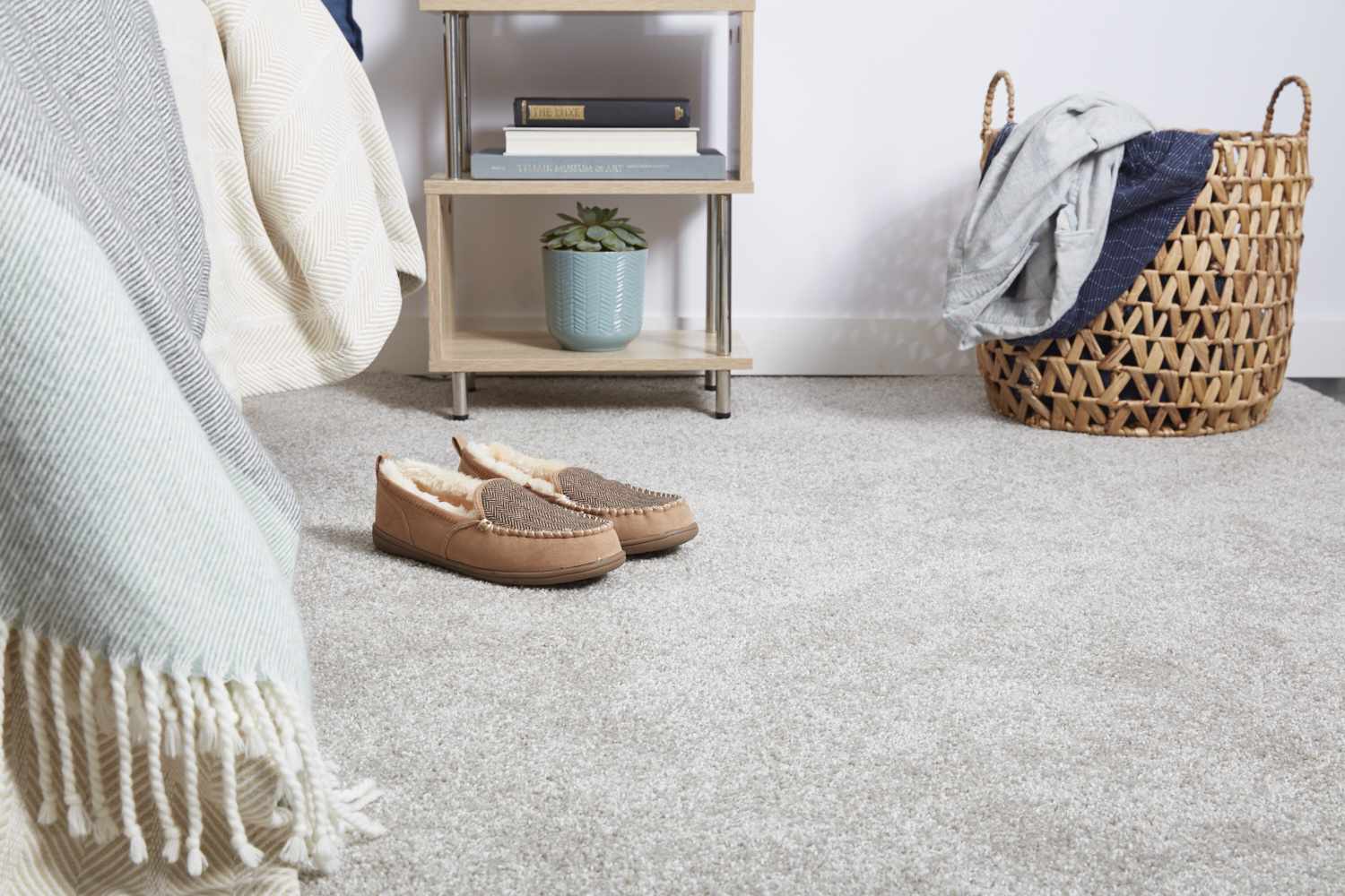 Piso de carpete com sapatos de lã ao lado da cama e cesto de tecido, em frente à mesinha de cabeceira