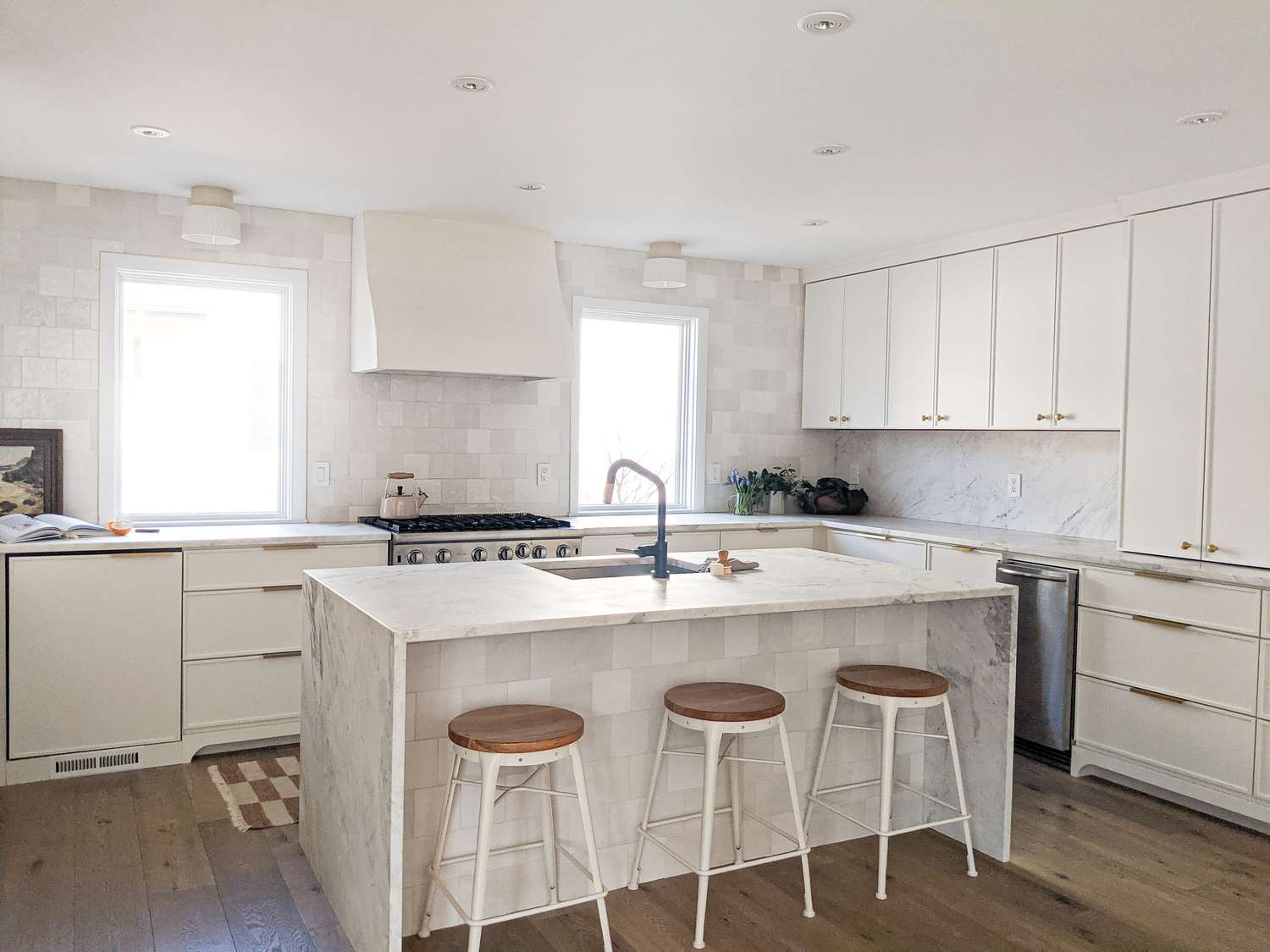 A cozinha branca na casa de Molly & Fritz em Shaker Heights apresenta uma ilha de mármore em cascata, ferragens pretas e janelas em ambos os lados do fogão
