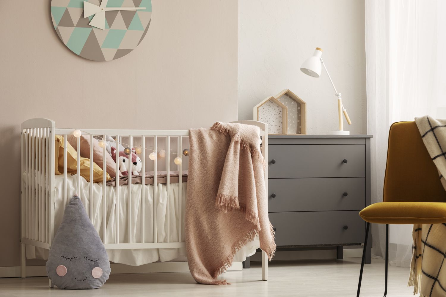 Travesseiros e brinquedos em um berço de madeira branco com manta rosa pastel em um quarto de bebê bem iluminado