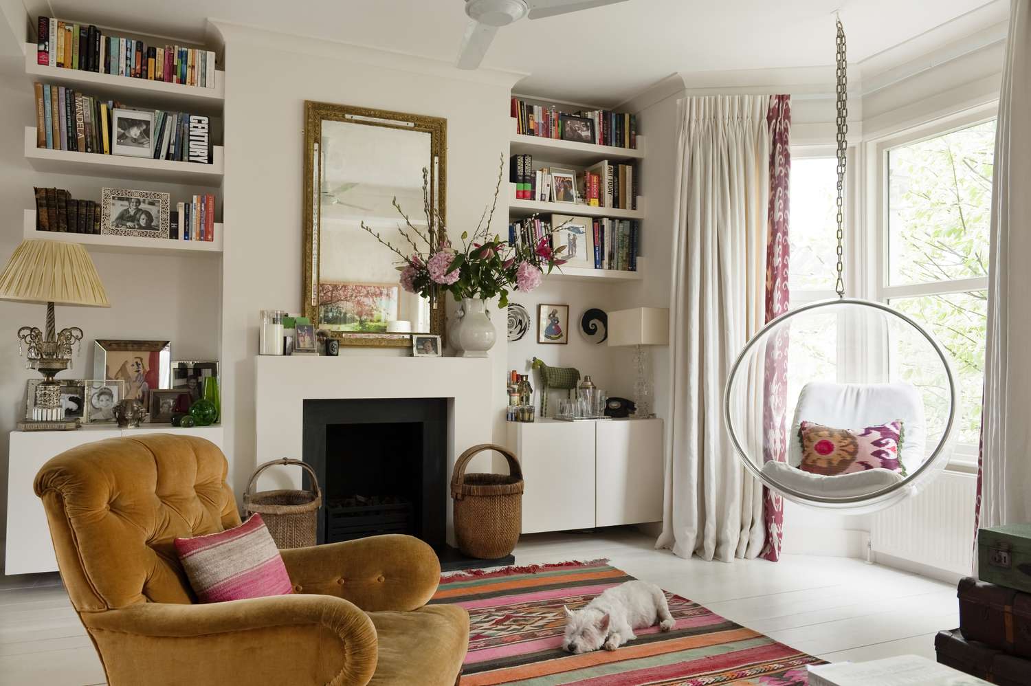 Salon avec une baie vitrée, des bibliothèques intégrées, un tapis de style aztèque, un fauteuil en velours jaune et un chien blanc qui se prélasse.