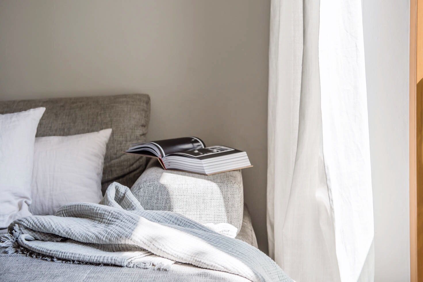 Image d'un canapé avec des coussins, une couverture et un livre ouvert sur le bord. Le canapé est à côté d'un rideau blanc