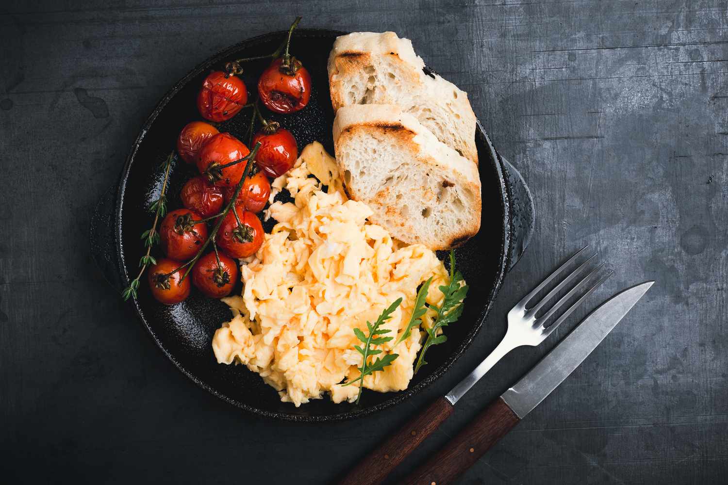 Assiette de petit-déjeuner noire contenant des œufs, des toasts et des tomates cerises rôties photographiée du dessus
