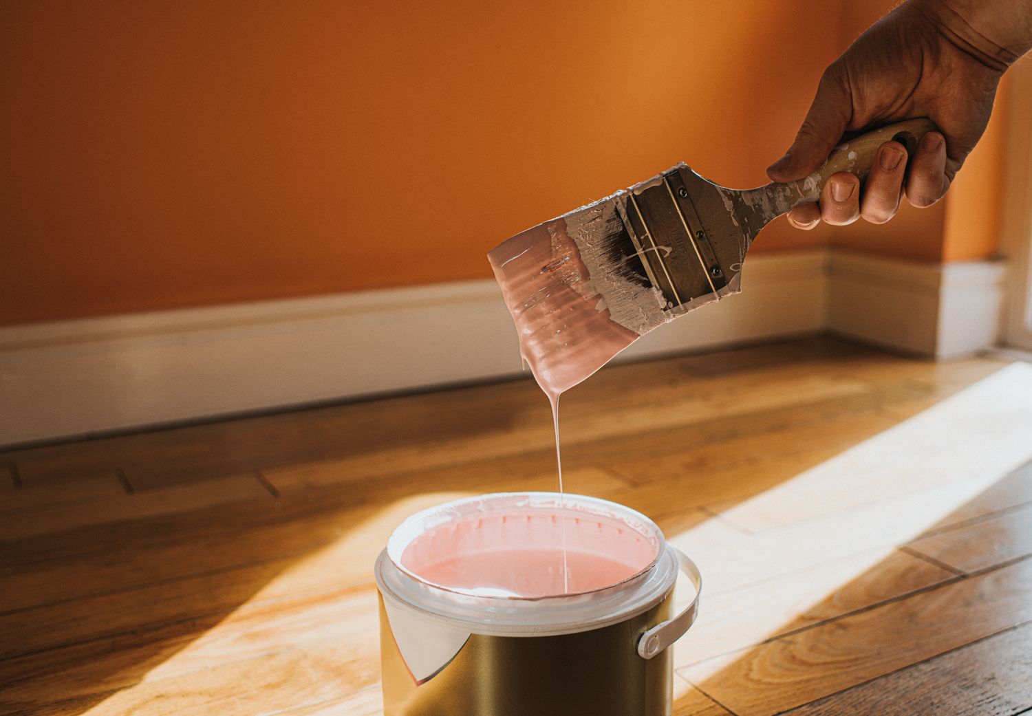Eine Hand taucht einen Pinsel in eine große Dose mit rosa Farbe, die zurück in den Topf tropft