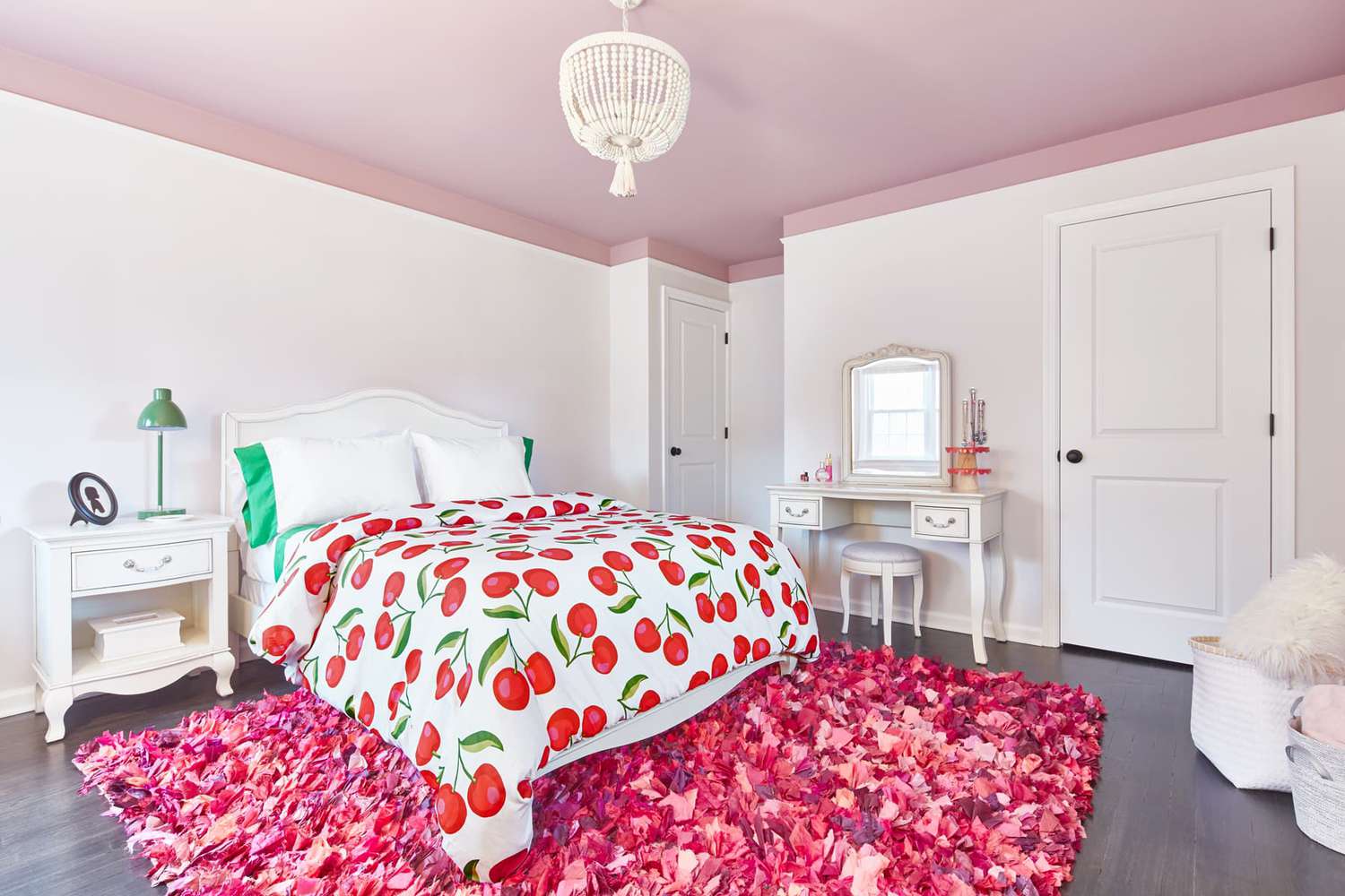 dormitorio con paredes blancas, una colcha con estampado de cerezas, almohadas verdes brillantes y lámpara de mesilla, 