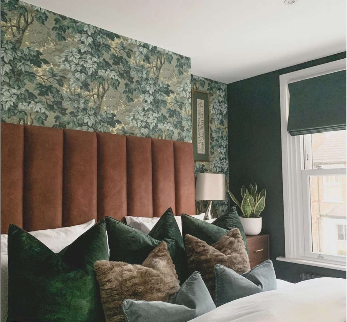 quarto com cabeceira de almofada vermelho-alaranjado queimado, papel de parede floral azul-esverdeado, almofadas verdes e azuis na cama, planta cobra no canto