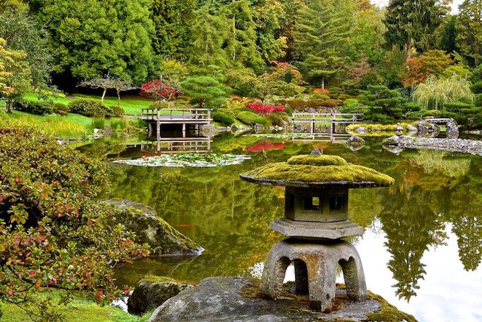 Großer Teich im japanischen Garten mit moosbewachsener Steinpagodenskulptur und Stein- und Holzbrücken im Hintergrund.