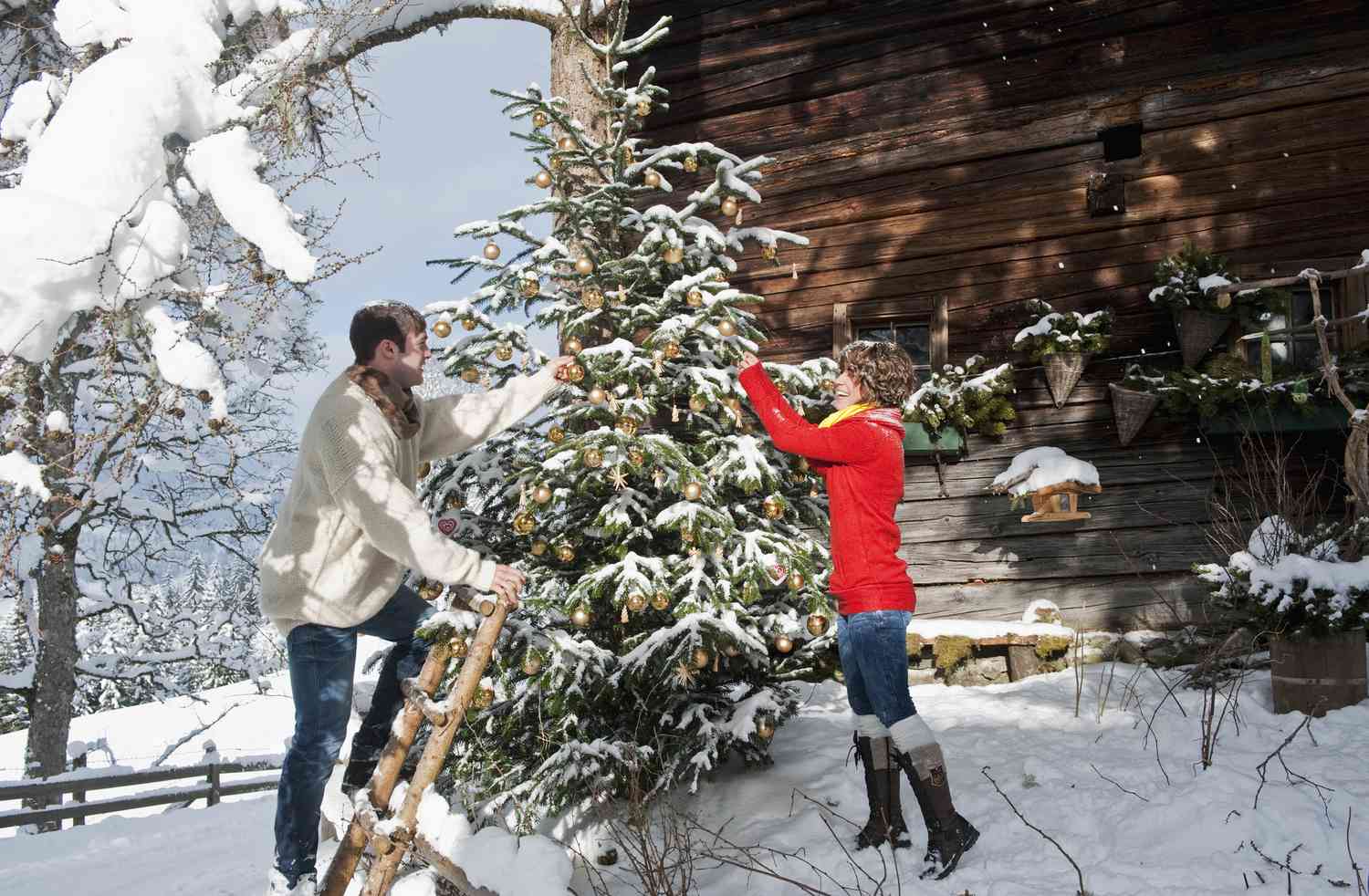 Paar mit einer Leiter beim Schmücken eines Weihnachtsbaums im Freien in einer verschneiten Landschaft.