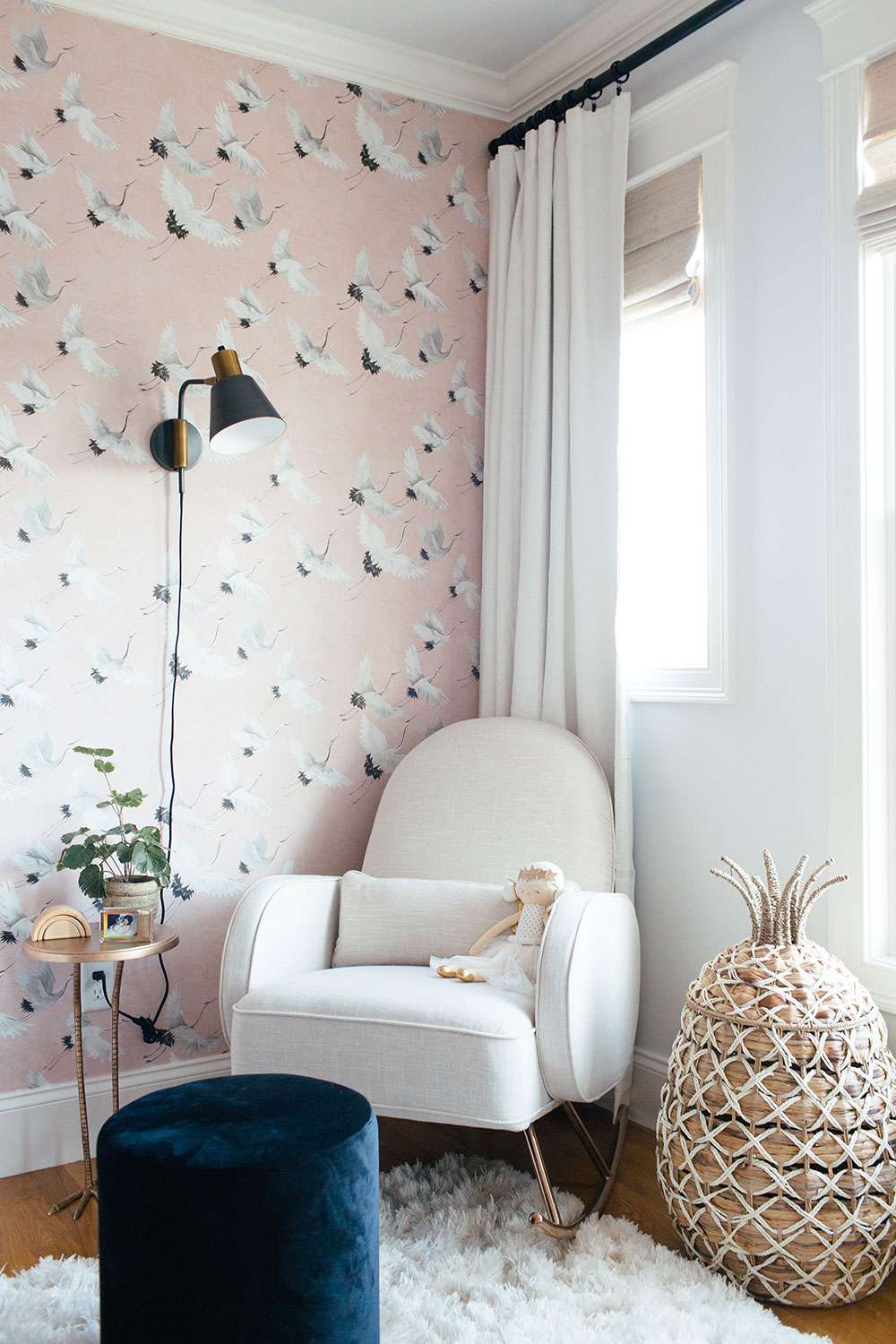 nursery with pink crane wall accent wall, black sconce, white rocking chair with wicker pineapple basket