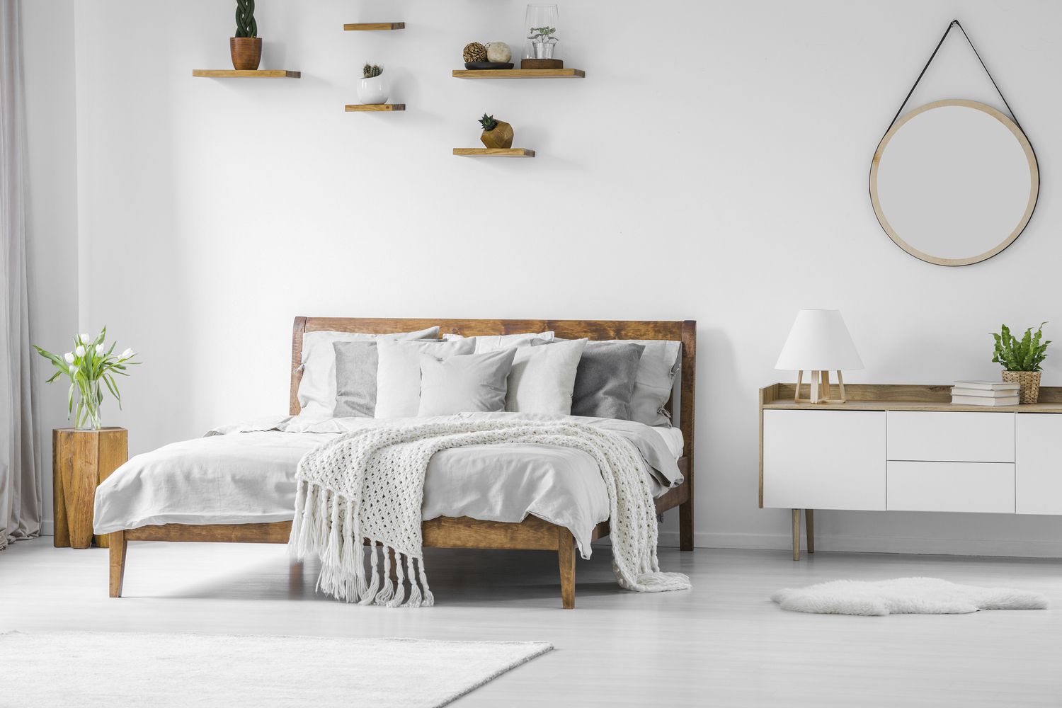 Grand lit confortable en bois avec linge, oreillers et couverture, table de nuit à côté et miroir rond suspendu sur un mur blanc dans un intérieur de chambre à coucher lumineux.