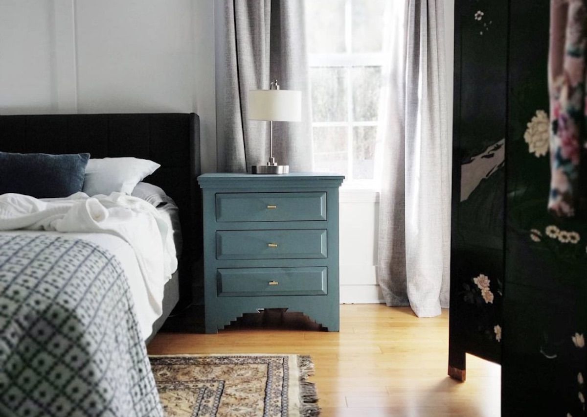 quarto com piso de madeira nua, mesa lateral azul-petróleo, roupa de cama azul, tapete de padrão de brechó no chão, paredes brancas