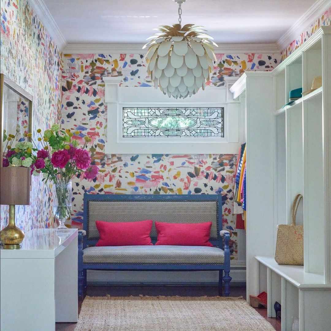 Un mudroom con colorido papel pintado con motivos de acuarela, un banco tapizado con brillantes cojines rosas y una gran lámpara de araña contemporánea blanca.