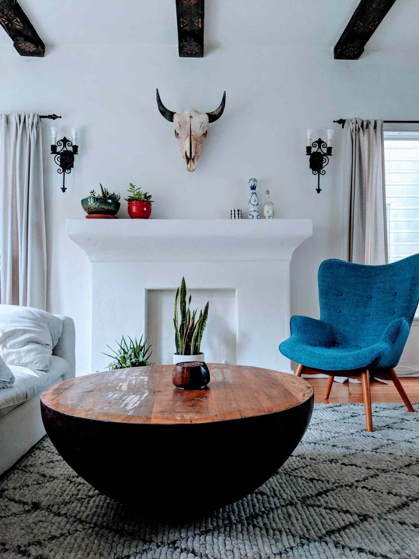 Salón boho chic con mesa circular, silla azul, chimenea de imitación y cabeza de toro en la pared.