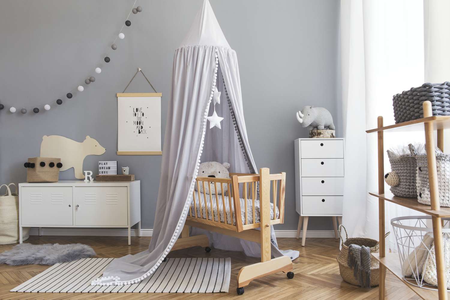 Stilvolle skandinavische Neugeborenenzimmereinrichtung mit Poster, weißen Möbeln, natürlichen Spielsachen, grauem Baldachin mit Sternen und Teddybären. Minimalistische und gemütliche Einrichtung des Kinderzimmers.