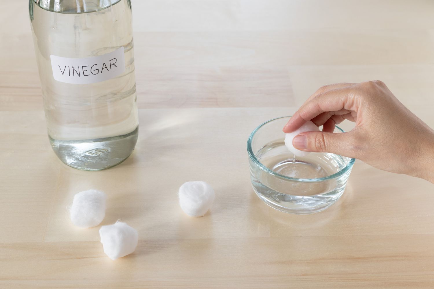 Distilled white vinegar glass bottle next to glass bowl with cotton balls being soaked