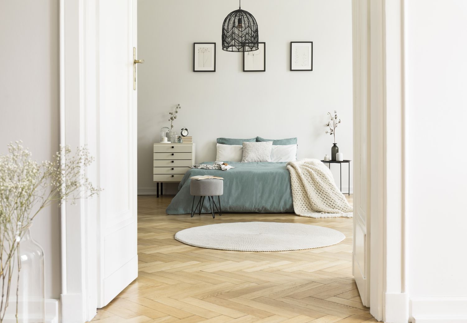 Foto real do interior de um quarto branco com tapete redondo, cama king-size com travesseiros e lençóis em tons pastéis, pôsteres simples e parquet em espinha de peixe