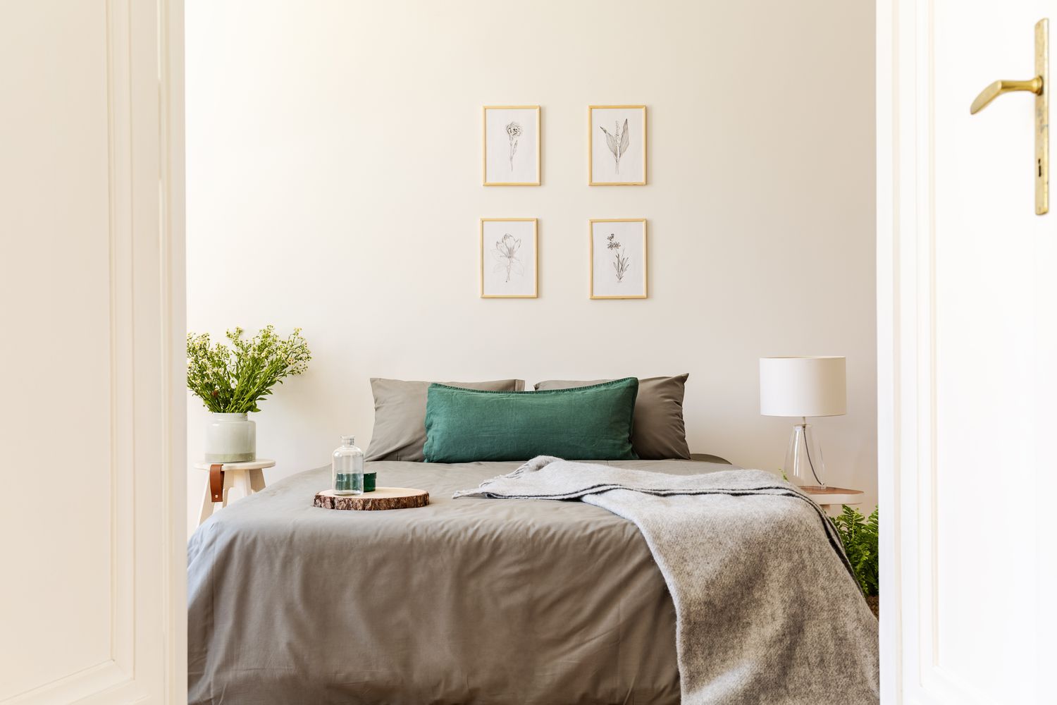 Ein Blick durch eine offene Tür in ein natürliches, sonniges Schlafzimmer mit grauen und grünen Laken und Kissen auf einem Doppelbett. Naturzeichnungen an der Vanille-Wand. Echtes Foto.