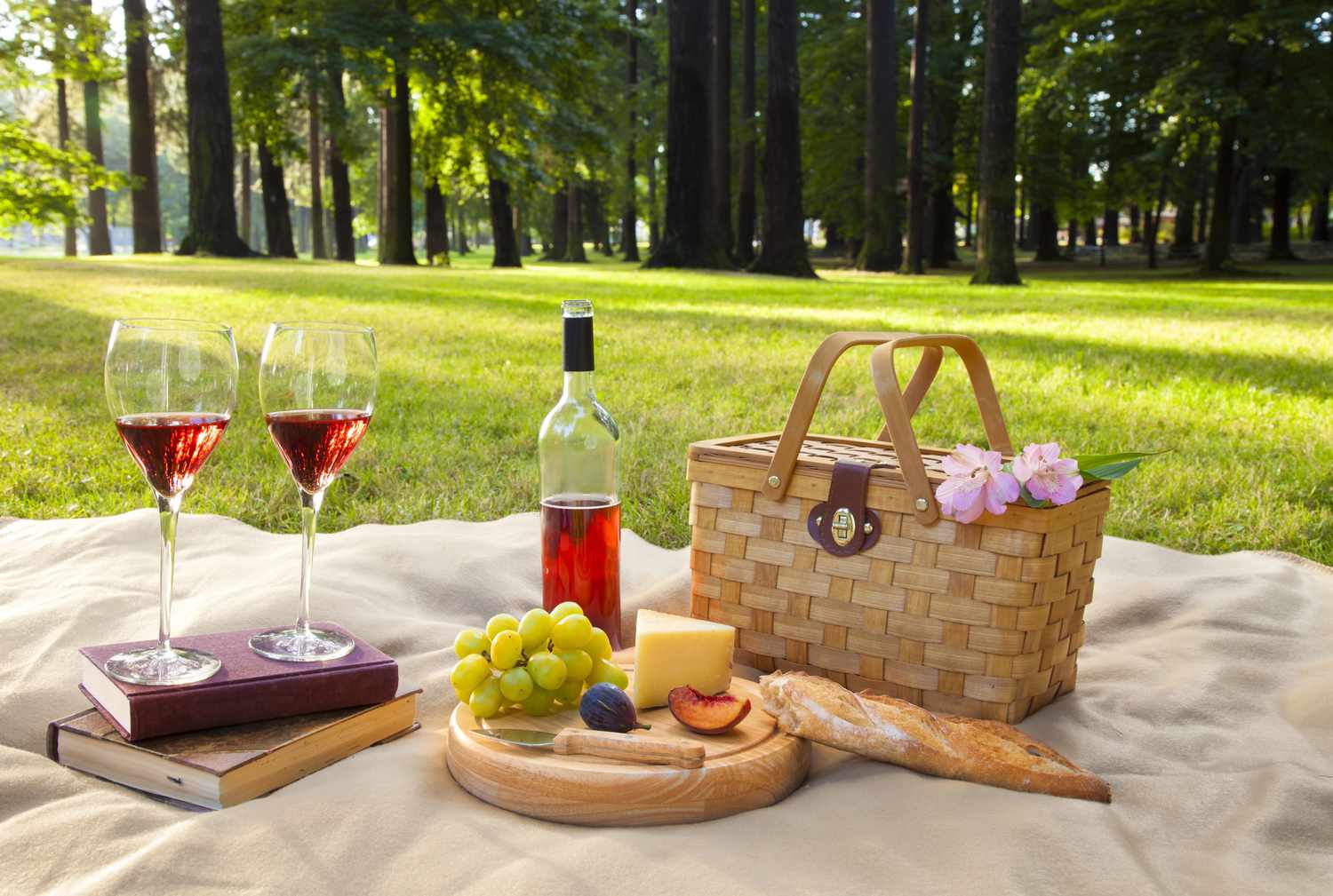 Vin, fromage, fruits, pain, et un panier de pique-nique sur une couverture sur l'herbe avec des arbres en arrière-plan