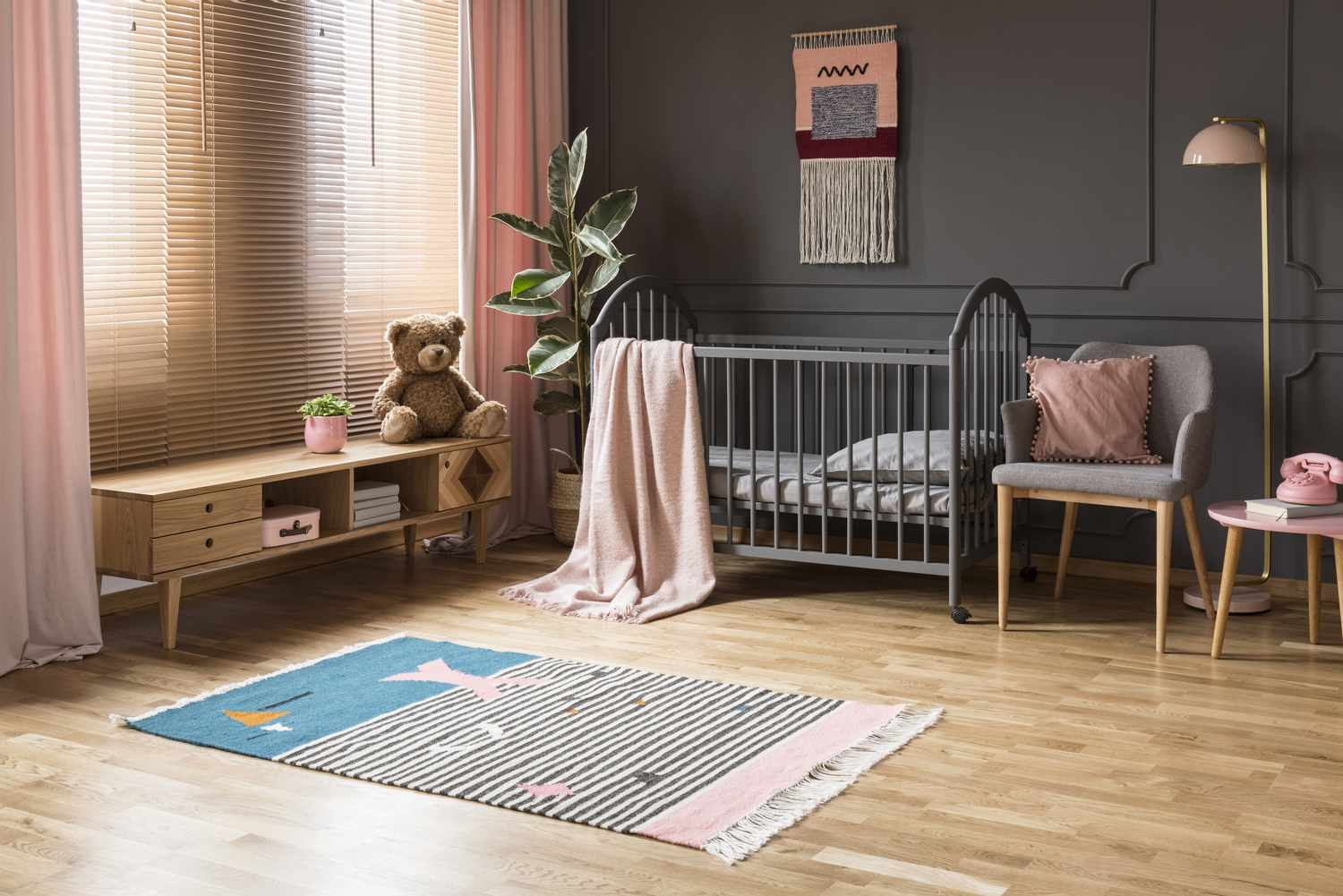 Echtes Foto eines Babybettchens, das zwischen einem niedrigen Schrank und einem Sessel, einer Lampe und einem Hocker in einem Kinderzimmer mit Holzboden und grauen Wänden mit Leisten steht