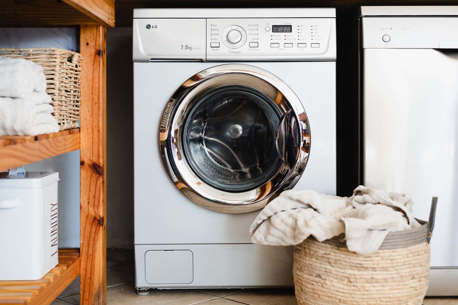 Máquina de lavar roupa ao lado de cesto com uma pilha de toalhas e prateleiras de madeira com materiais de lavanderia