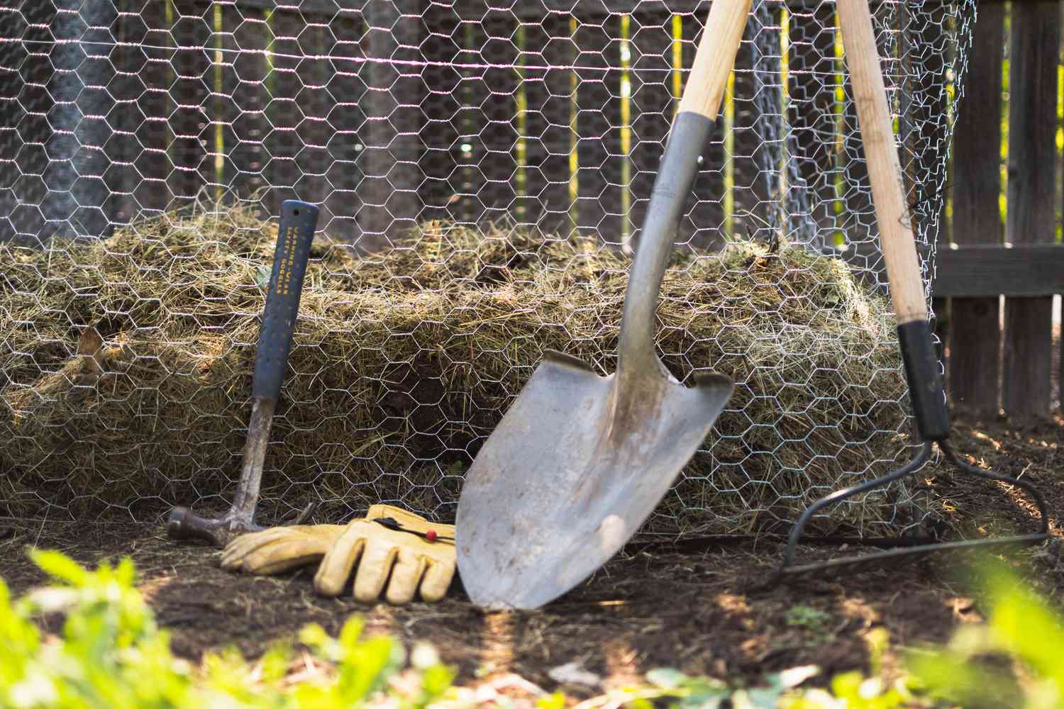 Selbstgebauter Komposter im Drahtzaun neben Schaufel, Harke, Gartenhandschuhen und Hammer im Hinterhof