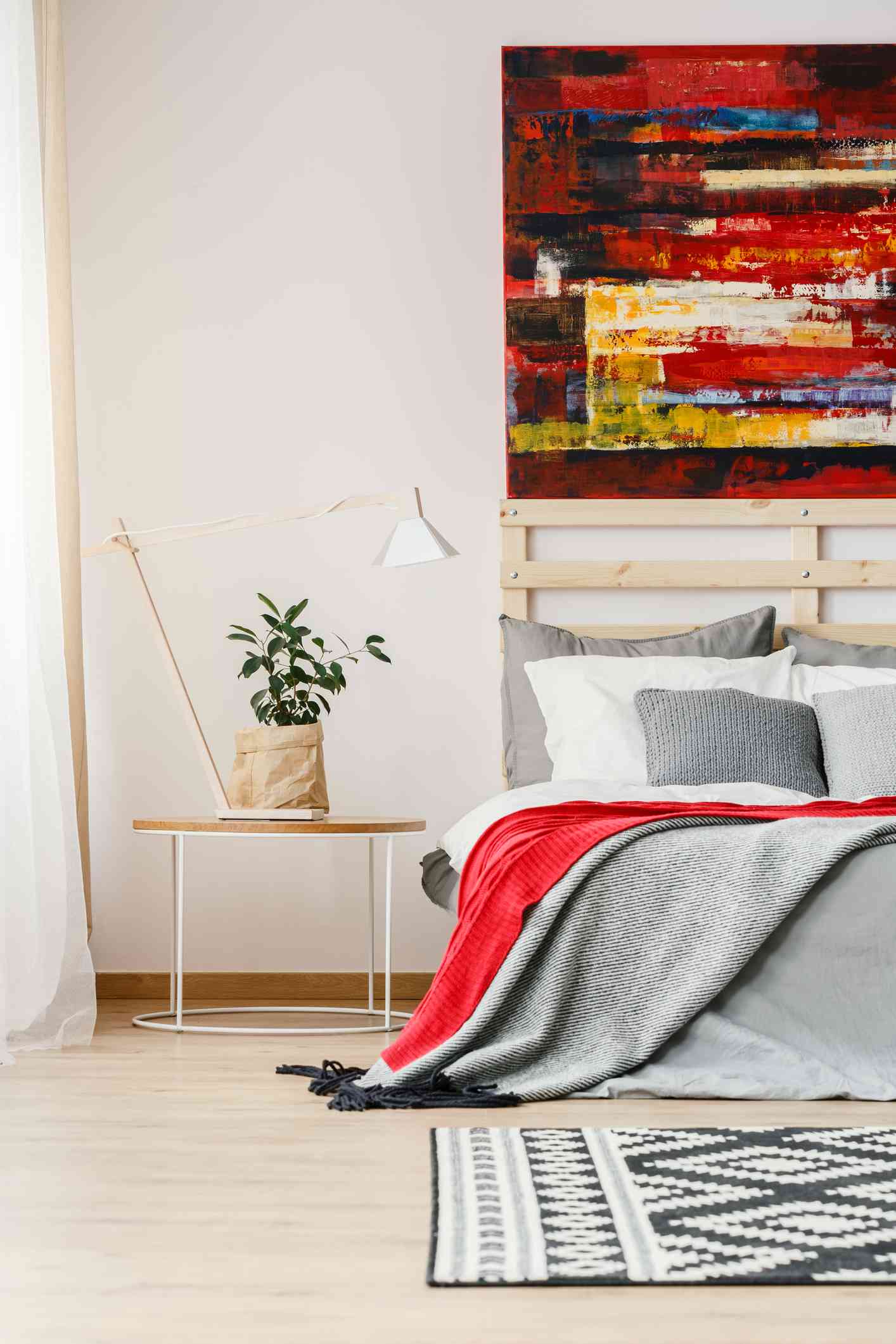 Manta gris y roja sobre cama junto a mesa con lámpara y planta en interior de dormitorio con cuadro.