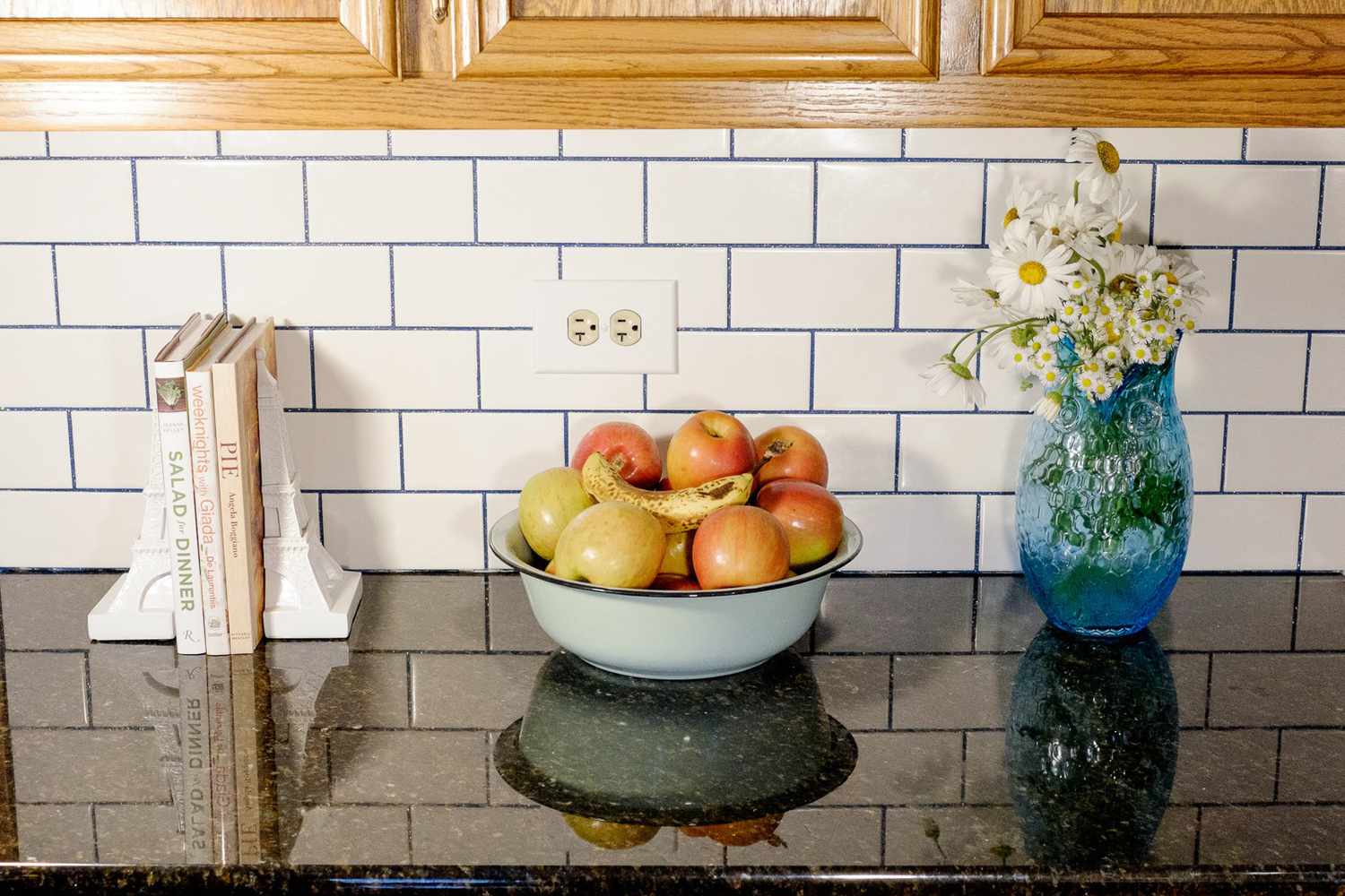 Livres, vase de fleurs, et bol de fruits sur le comptoir de la cuisine avec carrelage de métro blanc et coulis bleu étincelant