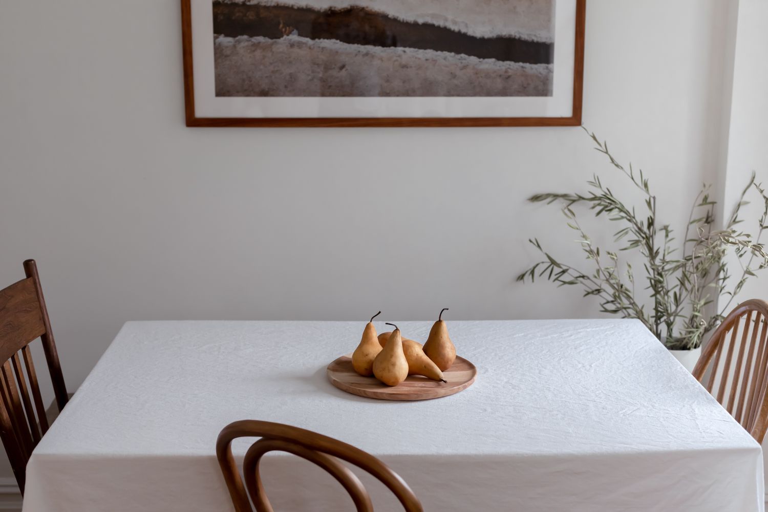 Mesa de comedor con mantel blanco y fuente de madera con peras junto a sillas de madera
