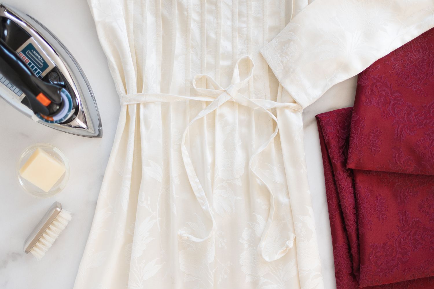 Weißes Damastkleid neben rotem Damast, gefalteter Stoff neben Bügeleisen und Reinigungsmaterial