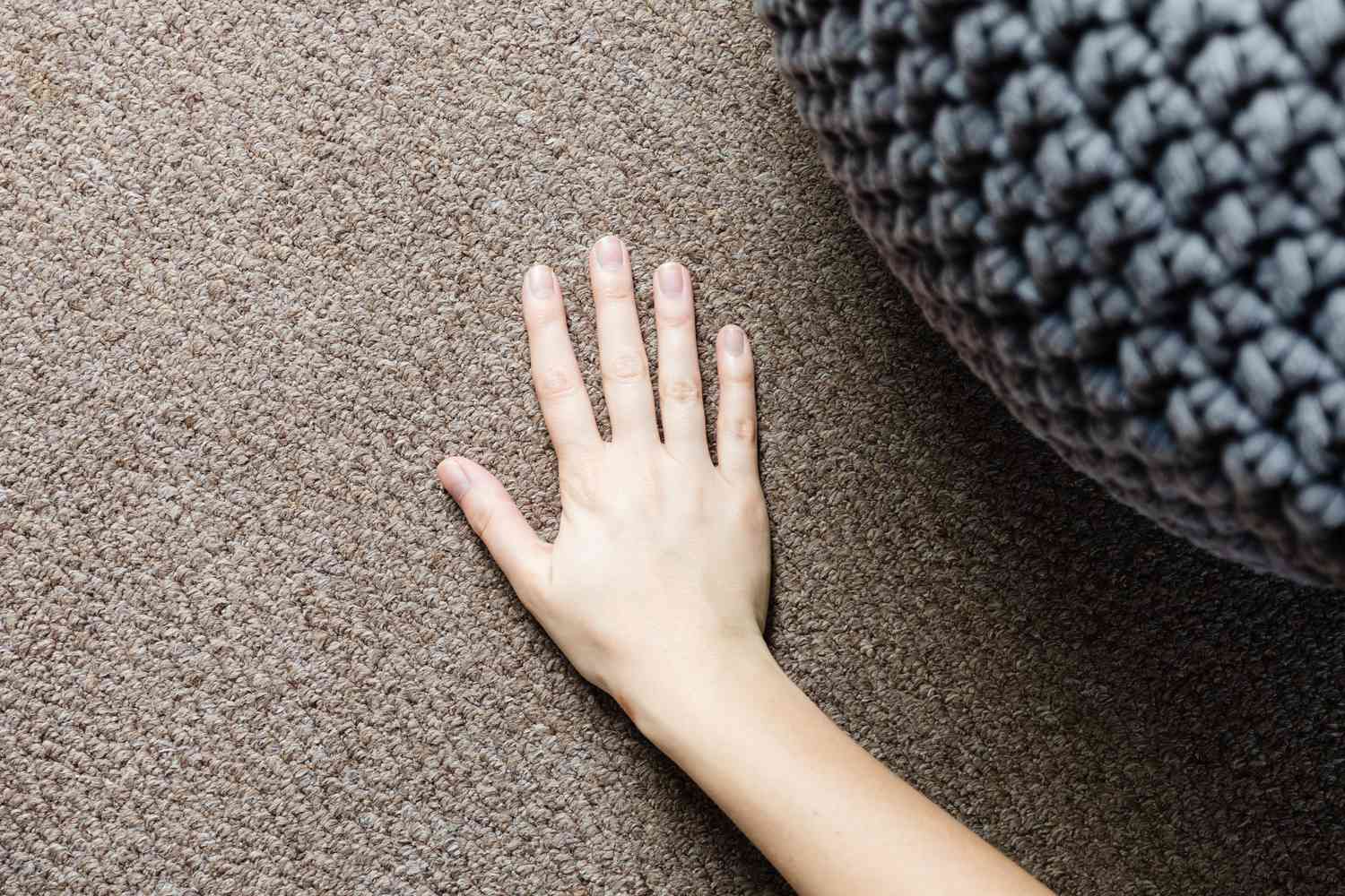 Hand passing over tan carpetted floor next to gray pouf