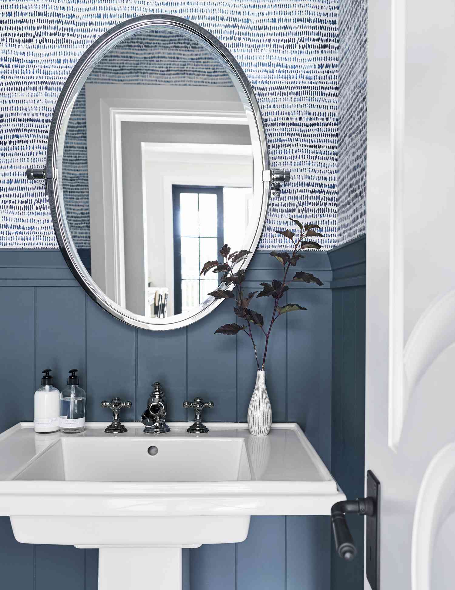 Lavabo de pedestal blanco con un espejo ovalado, papel pintado azul y blanco y revestimiento de madera azul pizarra