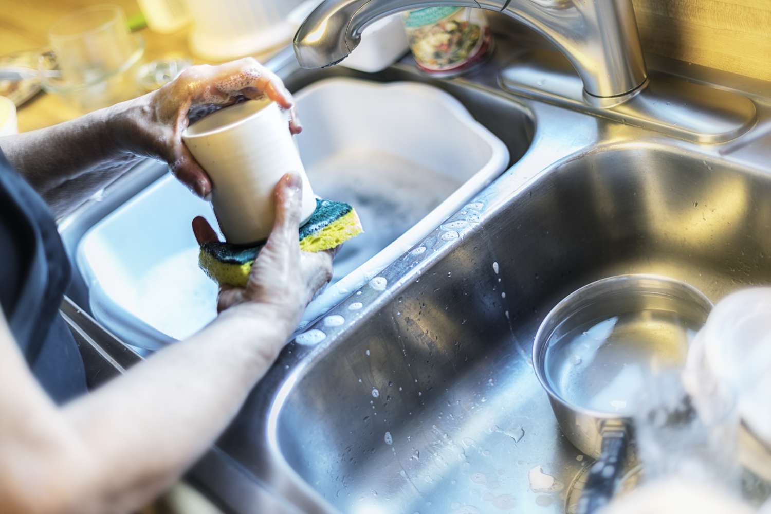 Eine Frau wäscht mit einem gelben Reinigungsschwamm eine Kaffeetasse und anderes Geschirr, Gläser und Kochtöpfe von Hand. Sie arbeitet in einer zweiteiligen Edelstahlspüle in der Küche.
