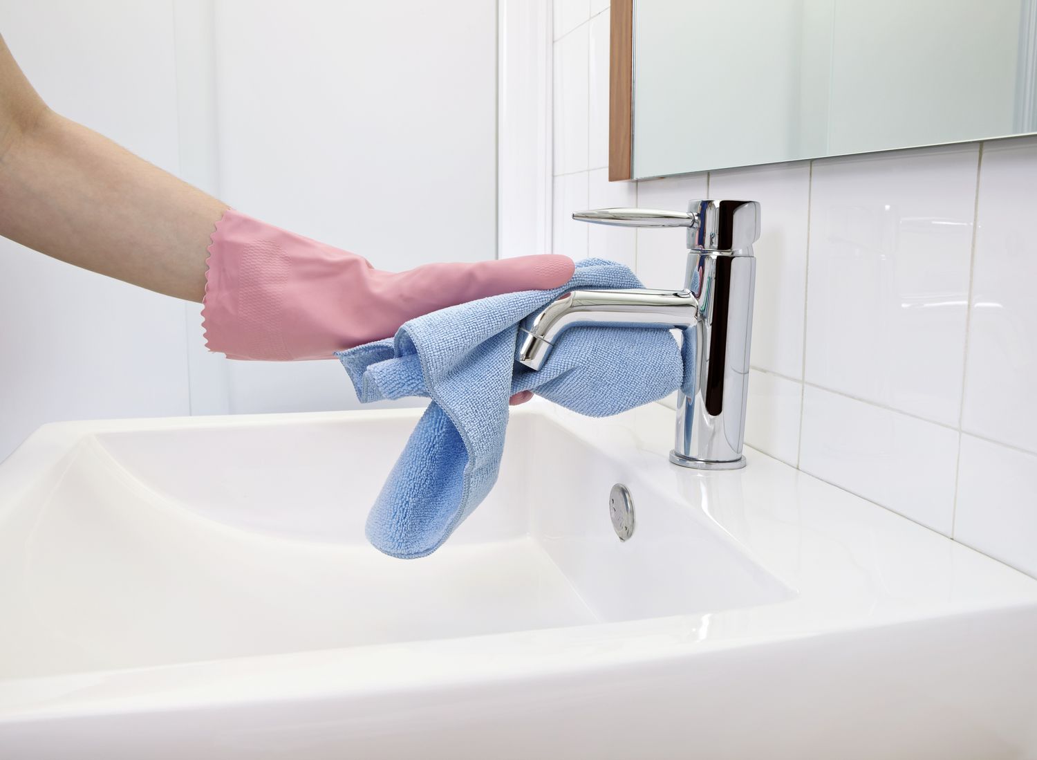 Mano femenina con guante de goma rosa limpiando grifo cromado del lavabo del baño con paño de microfibra azul