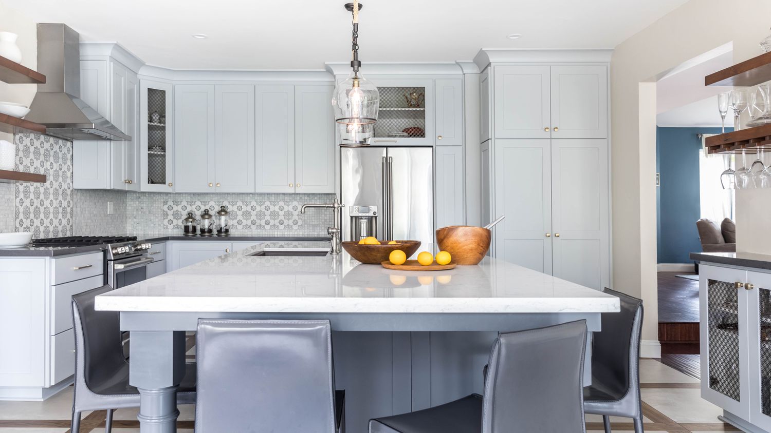 Armarios shaker de color gris azulado claro cubren una cocina que también tiene azulejos backsplash detallados en gris y blanco y suelos de madera enmarcados en piezas grises