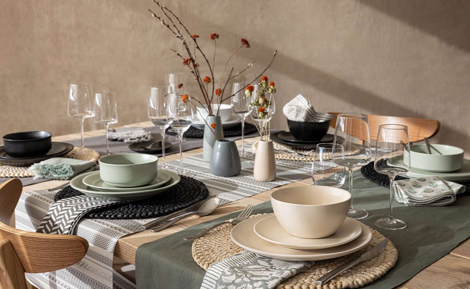 Un paysage de table d'assiettes beiges, noires et blanches sur différentes couches de napperons et trois vases de fleurs en céramique dans des couleurs similaires sont assis dans le centre de table