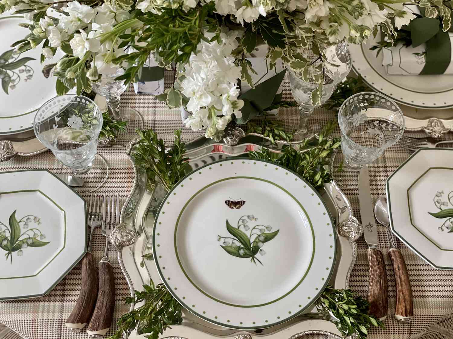 Une nappe à carreaux recouvre une table sur laquelle sont posées de délicates assiettes en céramique couvertes d'images de fleurs à feuilles. Le centre de table est composé de fleurs à feuilles blanches assorties aux assiettes