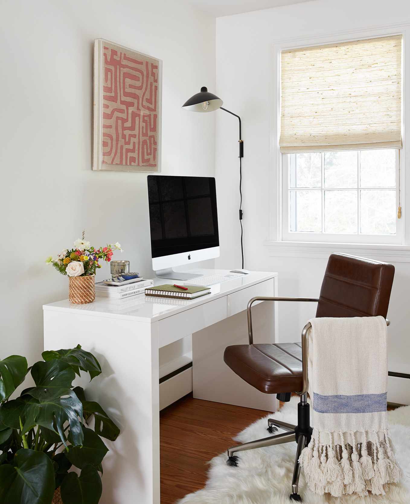 Uma mesa de escritório contemporânea branca abriga um computador apple abaixo de uma obra de arte emoldurada na cor pêssego