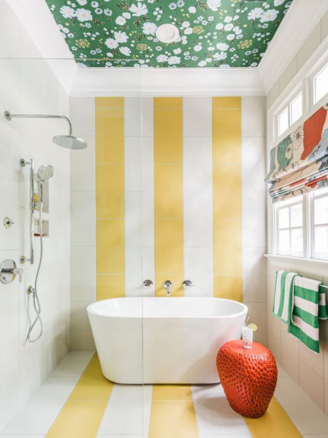 Baño a rayas amarillas y blancas con bañera exenta, techo de papel pintado floral verde y taburete de cerámica naranja